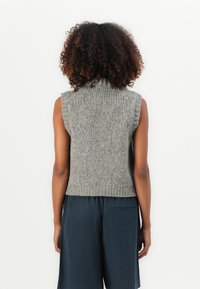 Persoon met krullend haar, gekleed in een grijze gebreide mouwloze trui en losse zwarte broek, staand tegen een plain witte achtergrond.