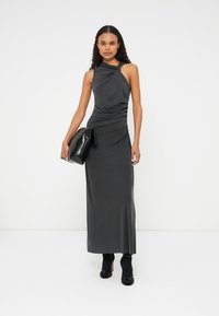 Crna haljina bez rukava s uvrnutim dekolteom, nabranim dizajnom i lepršavim siluetom, uparena s sjajnom crnom clutch torbom i cipelama s potpeticama.