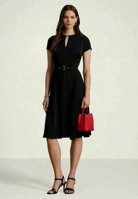 Moteris vilkinti juodą su diržu iki kelių suknelę ir juodus aukštakulnius sandalų, laikanti mažą raudoną rankinę, stovinti prieš paprastą foną.