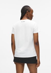 T-shirt bianco in cotone con maniche corte e colletto rotondo, caratterizzato da un piccolo logo nero al centro sul retro, indossato con una gonna nera.