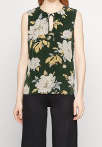 Ärmlös mörkgrön topp med stort vitt och gult blommotiv, nyckelhålsringning med knytning, bärs med svarta byxor.