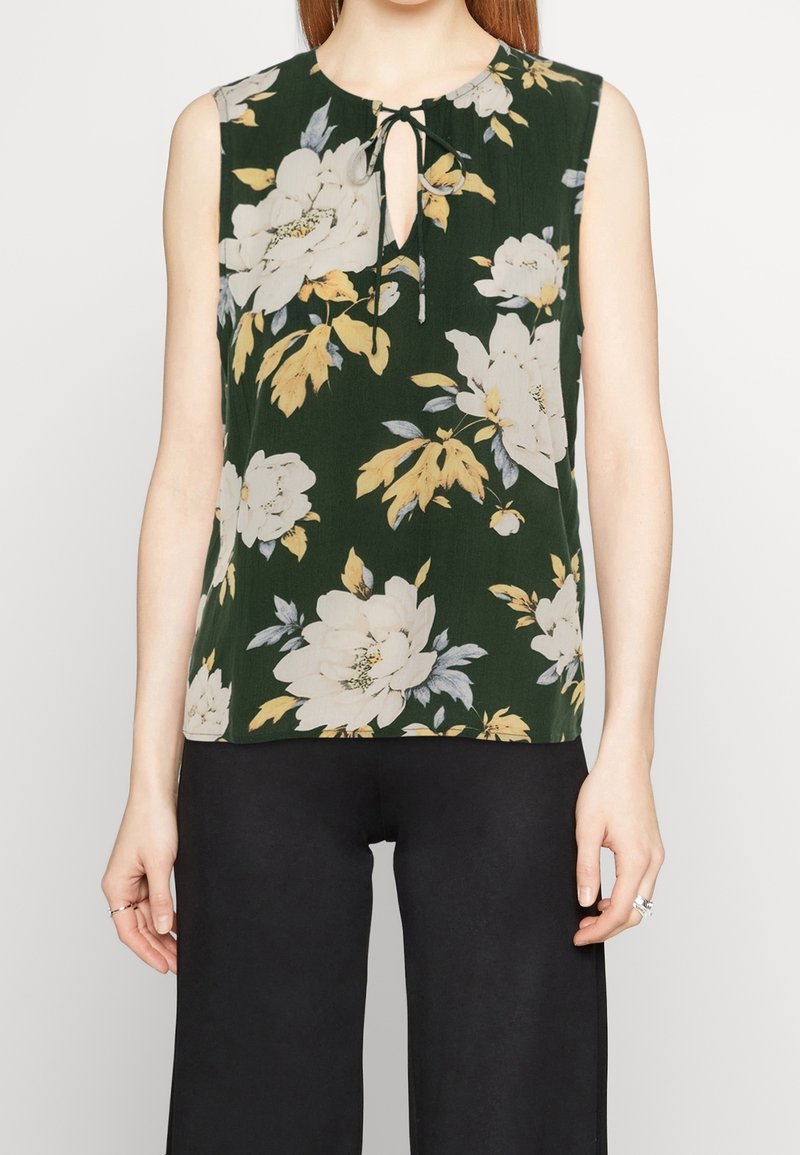 Ärmlös mörkgrön topp med stort vitt och gult blommotiv, nyckelhålsringning med knytning, bärs med svarta byxor.