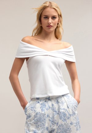 Weißes Off-Shoulder-Top mit drapiertem Stoff und figurbetontem Schnitt. Kombiniert mit plissierten hellblauen Hosen im floralen Muster.