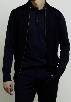 Mężczyzna ubrany w ciemnogranatową koszulkę polo z długim rękawem, dopasowaną kamizelkę zapinaną na zamek i spodnie, stojący z jedną ręką w kieszeni na jasnym tle.