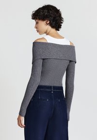 Frau mit kurzen lockigen Haaren, die einen grauen, gerippten Pullover mit schulterfreiem Schnitt über einem weißen Tanktop und hochtaillierten, dunkelblauen Jeans trägt und seitlich steht.