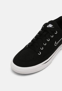 Svart canvas-sneaker med vita accenter, rund tå och texturerad gummisula. Har metallöglor och platta snören. Logotyp på tungan.