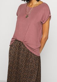 Roze kortemouwen top met opgerolde manchetten, knoop onderaan, gecombineerd met een zwarte rok met een klein oranje patroon. Gouden gelaagde ketting.