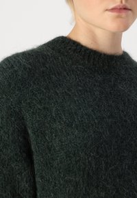 Σκούρο πράσινο πλεκτό πουλόβερ με στρογγυλή λαιμόκοψη, απαλή υφή και ελαφρώς χαλαρή εφαρμογή, το οποίο επιδεικνύει ένα φουντωτό σχέδιο νήματος.