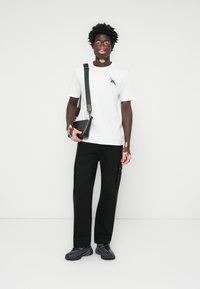 Weißes T-Shirt mit kurzen Ärmeln und Logo, schwarze Cargo-Hosen und schwarze Umhängetasche. Schwarze Sneakers mit strukturierter Detailierung. Einfarbiger Hintergrund.