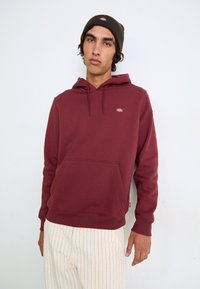 Hoodie bordeaux avec une poche avant, capuche arrondie à cordon de serrage et un petit patch logo. Associé à un pantalon crème rayé et un bonnet vert.