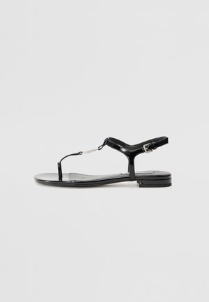 Sandalie plate din piele lacuită neagră, cu bretele între degete, curea la glezna și cataramă mică; textură netedă și design minimalist.
