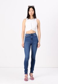 White twisted crop top, blue high-waisted skinny jeans, and purple strappy heels. Model stands against a plain white background.
