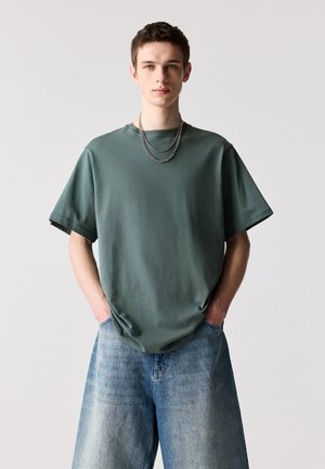 Tricou supradimensionat cu mâneci scurte, de culoare verde pal, realizat dintr-un material texturat, asortat cu blugi evazați de culoare albastru deschis, cu mâinile în buzunare.