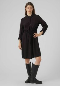 Rochie neagră cu nasturi, cu mâneci lungi, două buzunare pe piept, talie strânsă și material ușor, combinată cu cizme negre până la genunchi.