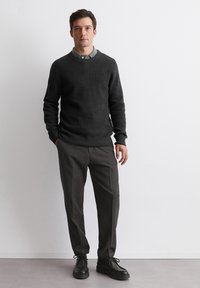 Dunkelgrauer gestrickter Pullover mit gerippten Bündchen, getragen über einem hellgrauen Hemd mit Kragen und kombiniert mit dunkelgrauen maßgeschneiderten Hosen und schwarzen Schuhen.