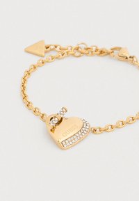 Βραχιόλι με αλυσίδα από χρυσό με μενταγιόν σε σχήμα καρδιάς, διακοσμημένο με διάφανα στρας και χαραγμένο με τη λέξη «GUESS»· διαθέτει κούμπωμα τύπου toggle.