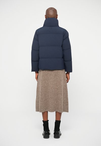 Tumesinine puhvjakken kõrge kraega, tikandiga, pikkade varrukatega, koos beeži tekstuuriga seeliku ja mustade pahkluu saabastega.