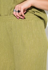 Olijfgroen stof met een textuurafwerking, voorzien van subtiele metallic draden. De tailleband is geraffineerd, wat het algehele casual ontwerp accentueert.
