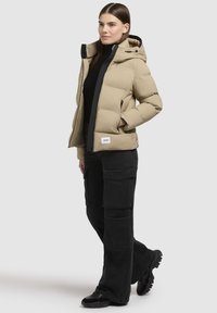 Beige Steppjacke mit Kapuze, ausgestattet mit einem Reißverschluss vorne und Seitentaschen. Kombiniert mit schwarzen Cargo-Hosen und schwarzem Schuhwerk.