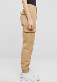 Khaki cargo kalhoty s bočními kapsami, zúženými jogger manžetami a hladkou bavlněnou texturou, doplněné bílými teniskami.