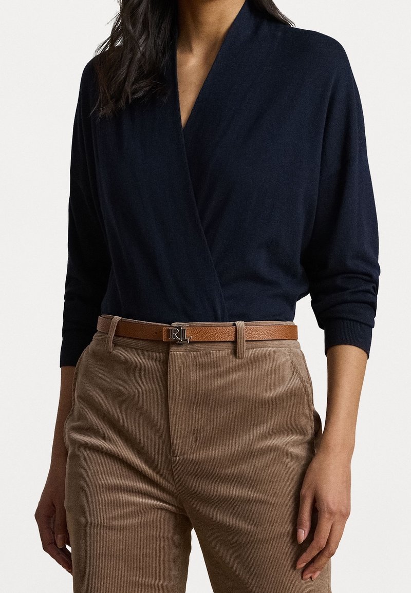 Femme portant un pull cache-cœur bleu marine, un pantalon en velours côtelé beige et une ceinture en cuir marron avec une boucle argentée RL, debout les mains détendues.