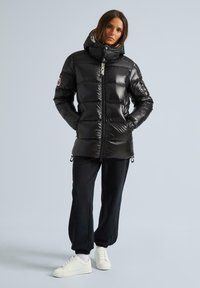 Zwarte pufferjack met een glanzende afwerking, hoge kraag en ritssluiting. Heeft elastische manchetten en zakken, gecombineerd met zwarte broek en witte sneakers.