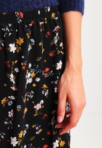 Jupe noire à motifs floraux colorés. Taille élastique et tissu léger. Gros plan sur une main reposant sur le tissu.