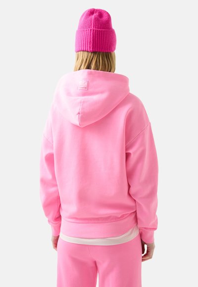 Rosa Kapuzenpullover mit lockerer Passform, gerippten Bündchen und Etikettendetail auf der Rückseite. Das Modell trägt eine passende rosa Strickmütze.