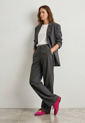 Frau steht mit den Händen in den Taschen, trägt einen grauen Anzug, ein weißes Hemd und leuchtend pinke Schnürschuhe vor einer schlichten grauen Wand.