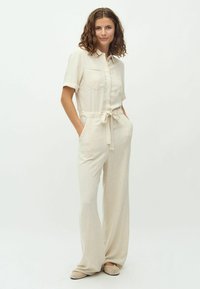 Jumpsuit de lino beige claro con mangas cortas, cintura ceñida con cinturón y dos bolsillos en el pecho. Diseño de pierna ancha con una textura suave.