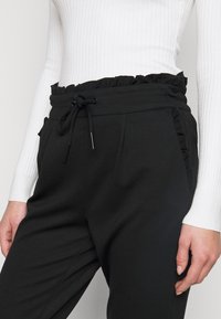 Calças de jogging pretas com cintura franzida, fecho com cordão e bolsos laterais. Material suave e textura lisa.