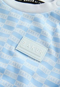 Svetlomodrá košeľa s krátkym rukávom s jemným károvaným vzorom a nápisom "BAKER" vytlačeným bielou farbou. Obsahuje textúrovanú modrú nášivku s logom.