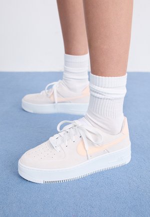 Jambes portant des chaussettes blanches montantes et des baskets blanches avec des logos Swoosh rose pâle, debout sur un tapis bleu.