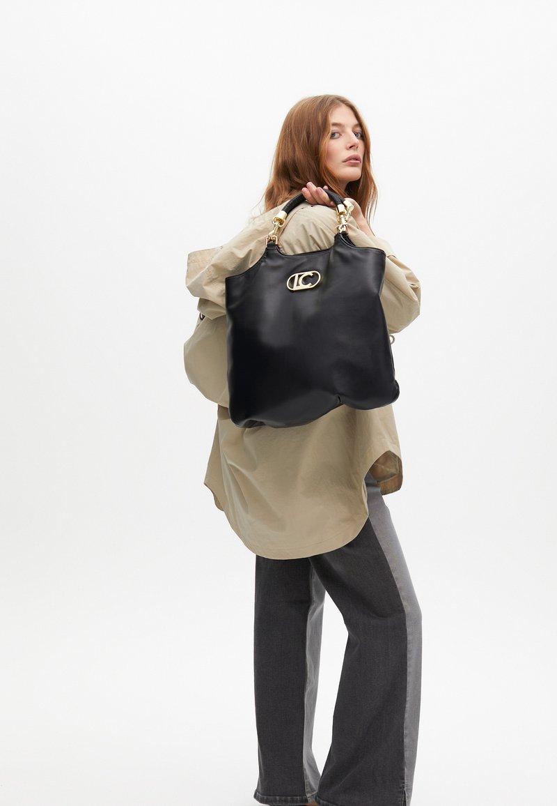 Modelka s dlouhými vlasy drží velkou černou kabelku se zlatým logem, má na sobě béžový oversize top a šedé džíny na bílém pozadí.