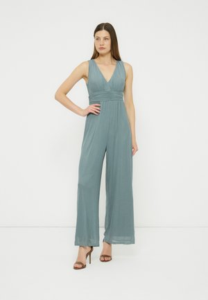 Vrouw staat met hand op heup, draagt een mouwloze stoffig blauwe wijde pijpen jumpsuit en bruine sandalen met hoge hakken tegen een witte achtergrond.