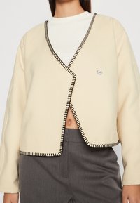 Kremowa krótka kurtka z czarnymi przeszyciami, o fasonie kopertowym, z długimi rękawami i małym, srebrnym guzikiem jako akcentem.