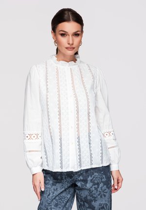 Femme portant un chemisier en dentelle blanche avec un col à volants et des manches longues, associé à un pantalon à motif floral foncé sur un fond uni.