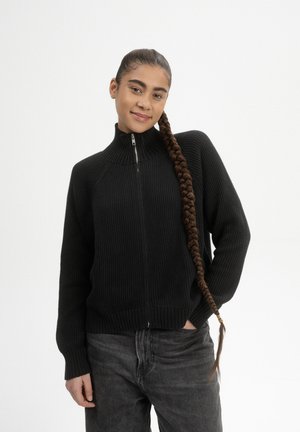Zwarte geribbelde ritstrui met hoge kraag, met lange mouwen en een aansluitende onderkant, gecombineerd met grijze jeans.