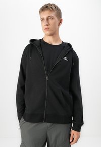 Joven con cabello rubio corto, vestido con una chaqueta con cremallera negra y pantalones grises, de pie contra un fondo blanco, mirando hacia un lado.