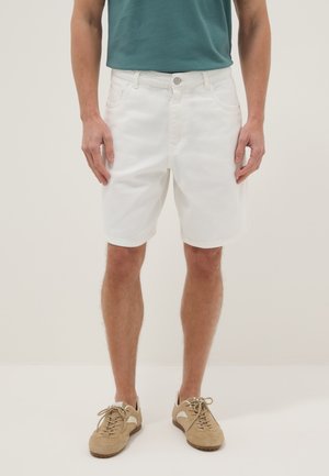 Uomo che indossa pantaloncini di jeans bianchi, t-shirt turchese e scarpe casual beige in camoscio con lacci, in piedi su uno sfondo chiaro e semplice.