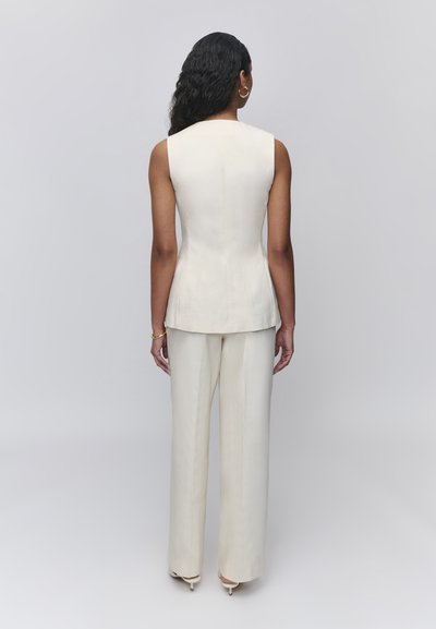 Femme aux longs cheveux noirs portant un gilet blanc sans manches sur mesure et un pantalon large assorti, debout devant un fond gris clair uni.