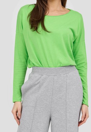 Nainen, joka pukeutunut kirkkaanvihreään pitkähihamaiseen paitaan, joka on tungettu korkeavyötäröisiin vaaleanharmaisiin housuihin, joissa on joustava vyötärönauha.