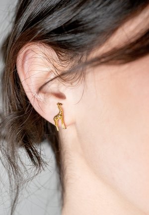 Gold giraffe-shaped earring with intricate detailing and a shiny finish, worn on a human ear, accented by dark hair.
