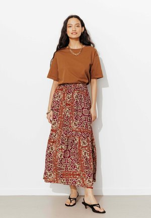 Femme portant un haut marron à manches courtes, une jupe longue à motifs rouges et beiges, des sandales noires à talons, un collier en chaîne dorée et un bracelet, debout contre un mur blanc.