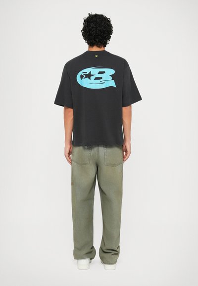 Schwarzes, übergroßes T-Shirt mit einem großen blauen Grafikdesign auf dem Rücken. Kombiniert mit ausgewaschenen grünen Jeans und weißen Sneakers.