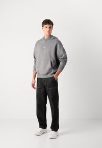 Grå hettegenser med frontlomme, sammen med svarte vide bukser. Modellen har på seg hvite sneakers, og viser en avslappet passform.