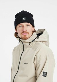 Giacca impermeabile beige con colletto alto e zip nera, indossata con un berretto in maglia nera con logo. Design semplice e funzionale.