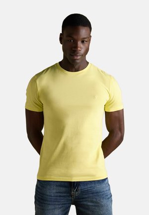 Mand står med hænderne bag ryggen iført en ensfarvet gul T-shirt med korte ærmer og blå jeans foran en hvid baggrund.