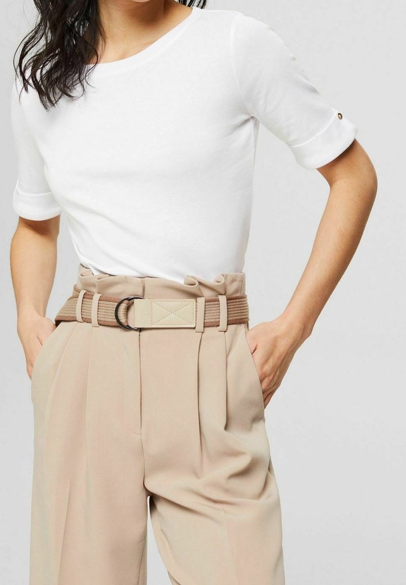 Femme portant un haut blanc à manches courtes rentré dans un pantalon beige plissé taille haute avec une ceinture en tissu assorti et les mains dans les poches.