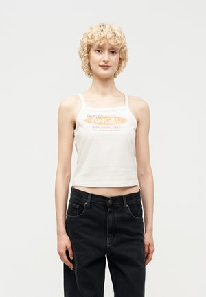 Młoda osoba o kręconych blond włosach, ubrana w biały krótki top na ramiączkach z napisem "Angel" oraz luźne czarne dżinsy, stojąca na tle gładkiego tła.